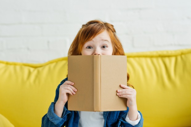 Kleines Mädchen lugt mit den Augen über den Rand ihres Buches und lächelt spitzbübisch in die Kamera | Credit: iStock.com/LightFieldStudios