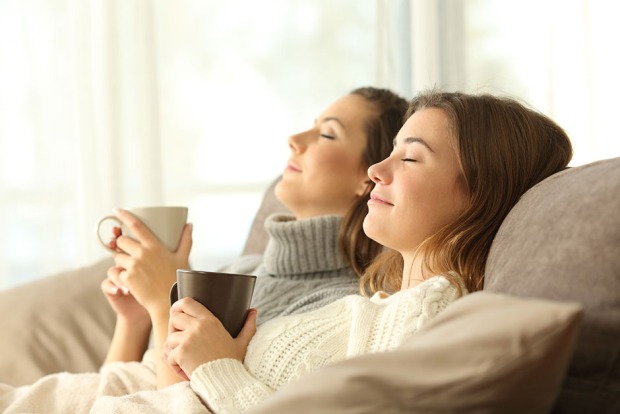 Zwei Freundinnen sitzen nebeneinander auf einer Couch mit geschlossenen Augen und träumen vor sich hin | Credit: iStock.com/AntonioGuillem