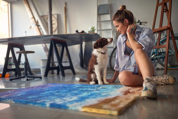 Junge Frau sitzt mit ihrem Hund vor ihrem selbstgemalten Bild | Credit: iStock.com/LuckyBusiness