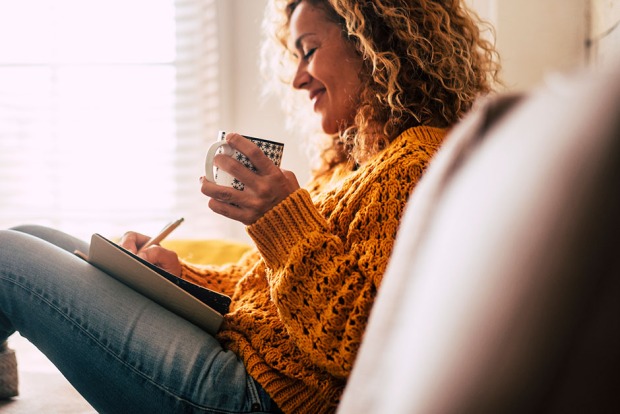 Eine glückliche Frau sitzt schreibend auf ihrer Couch | Credit: iStock.com/simonapilolla