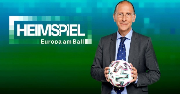 Peter Filzmaier vor einer Wand mit der Aufschrift: "Heimspiel. Europa am Ball." In der Hand hält er einen Fußball