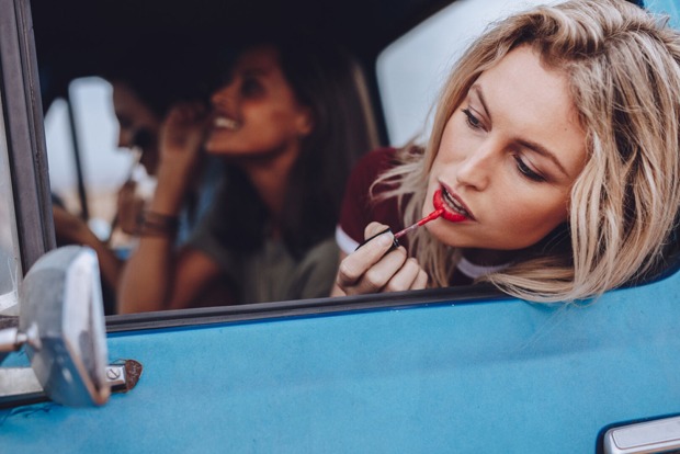 Junge Frau schminkt sich mit Blick in den Außenspiegel ihres Autos | Credit: iStock.com/jacoblund