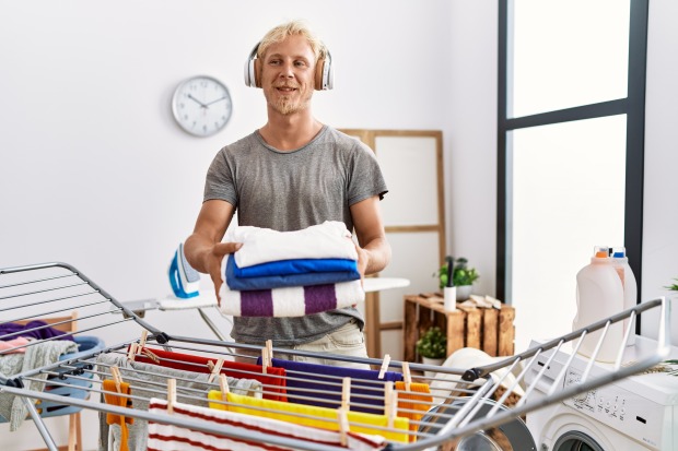 Mann faltet Wäsche vor einem Wäscheständer| Credit: iStock.com/AaronAmat