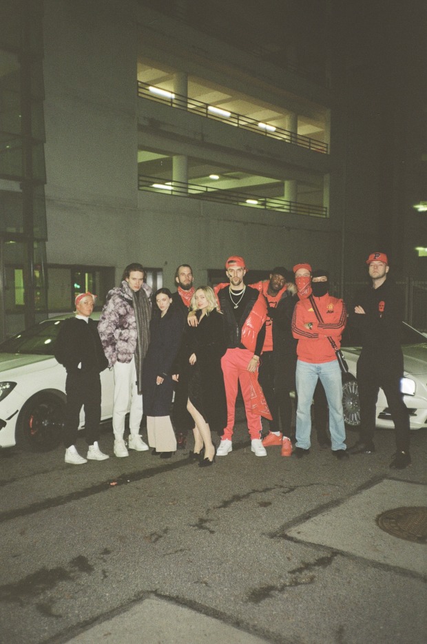 Eine Gruppe junger Männer und Frauen in Hip Hop-Outfits vor zwei Luxussportwägen und einem Gebäude bei Nacht