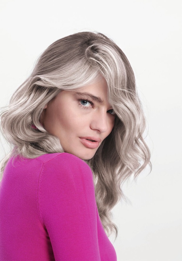 Frau mit blonden Haaren und weißen Strähnen vor einer weißen Fotowand mit einem pinken Pullover.