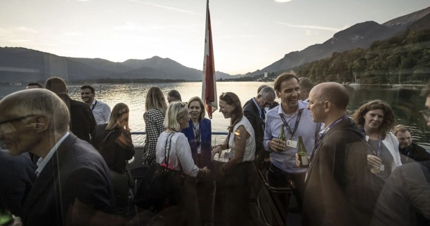 Grüppchen stehen am Schiffsdeck, im Hintergrund See und Berge