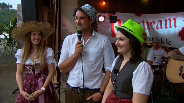 Zwei Frauen stehen neben einem Mann. Sie sind bei einem Fest und er spricht durch ein Mikrofon.