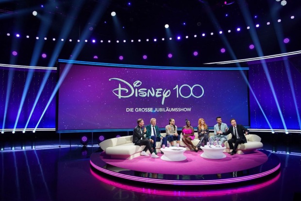 Prominente Gäste auf einer beigen Couch. Dahinter eine Leinwand auf der steht" Disney 100".
