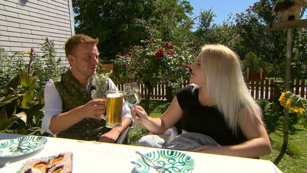 Bauer Johannes stößt mit Kandidatin Jenny an. Er trinkt ein Bier, sie einen Wein. Sie trägt offene blonde Haare und ein schwarzes Shirt, er eine grüne Tracht und ein weißes Hemd.