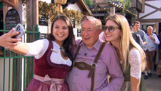 Richard Lugner trägt eine Tracht und ist umgeben von zwei jungen Frauen. Sie machen ein Selfie mit ihm.
