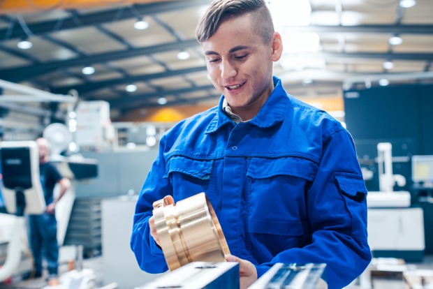 Ein junger Mann in einem blauen Overall, der in einer Fertigungshalle ein Bauteil inspiziert
