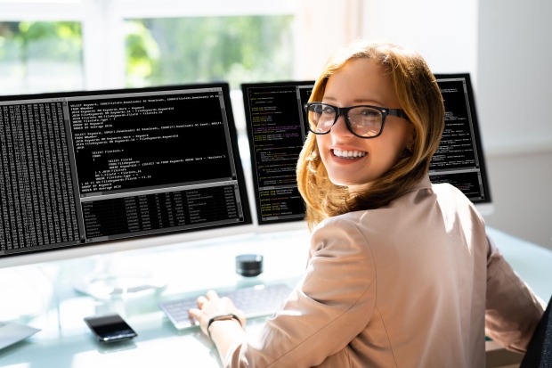 Eine junge Frau, die vor zwei Computermonitoren sitzt und in die Kamera lächelt