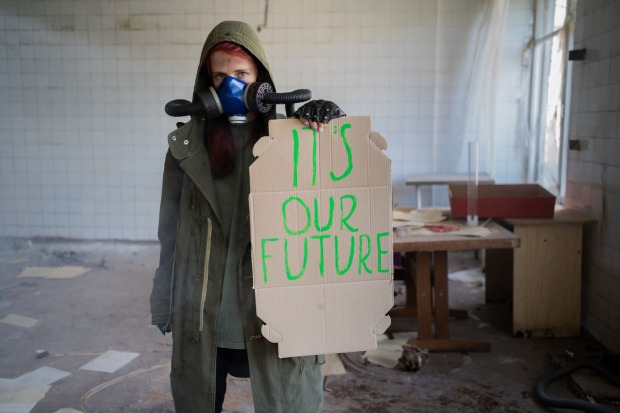 Ein weiblicher Prepper mit Gasmaske, der in einem heruntergekommenen Büroraum ein Schild mit der Aufschrift "It's our Future" in die Kamera hält