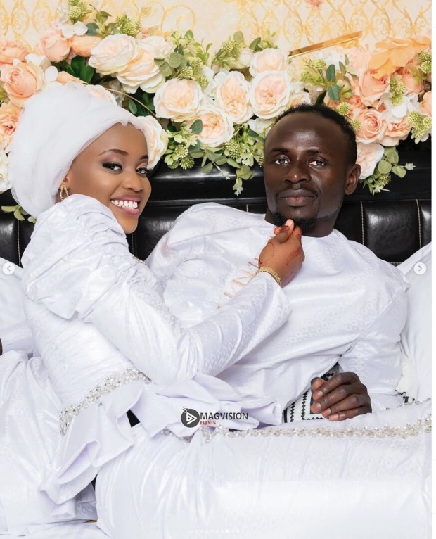 Sadio Mané und seine Ehefrau Aisha Tamba. Sie tragen weiße Roben und liegen ein einem Bett.