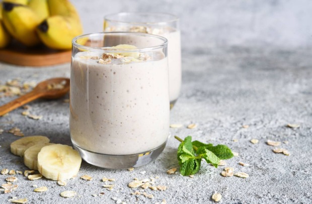 Zwei Gläser, gefüllt mit einem hellen Smoothie auf einer Steinplatte. Zur Dekoration liegen auf der Steinplatte 3 aufgeschnittene Bananenscheiben und ein kleiner Zweig mit Minzblättern.