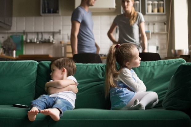 Zwei Kinder sitzen mit verschränkten Armen Rücken an Rücken auf dem Sofa.