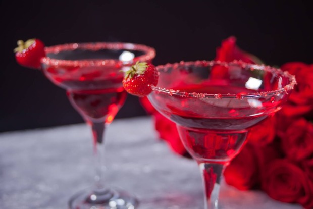 Zwei leuchtend rote, exotische alkoholische Getränke in transparenten Gläsern, begleitet von roten Rosen für ein romantisches Abendessen.
