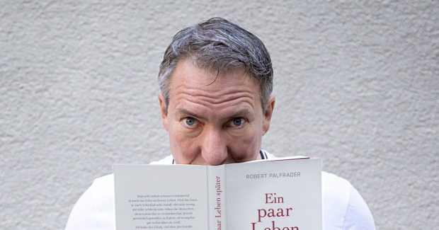 Robert Palfrader hält sein Buch "Ein paar Leben später" und schaut über den Rand hinweg.