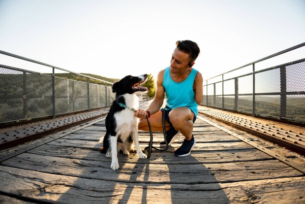 Junger Mann in türkisfarbenem T-Shirt und schwarzen Shorts, der in der Hocke seinen Border Collie streichelt.