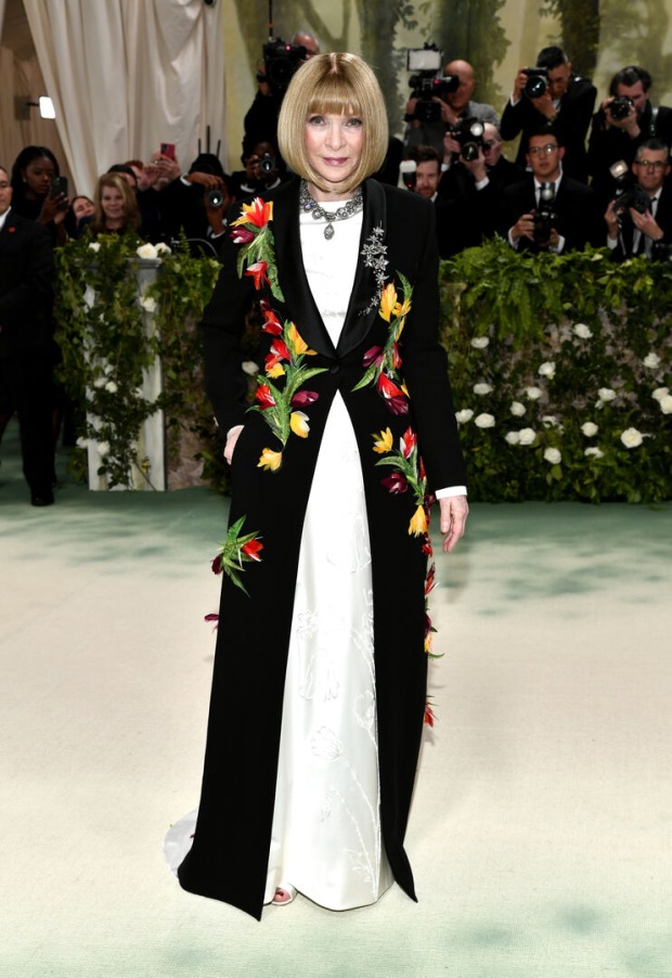 Eine Frau mit Bob in einem weißen Kleid, darüber trägt sie einen schwarzen Mantel mit Blumen