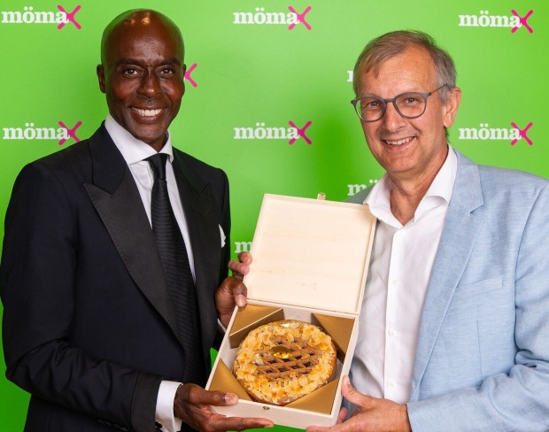 Bruce Darnell und Thomas Saliger vor einer "mömax"-Wand mit einer Linzer Torte in der Hand