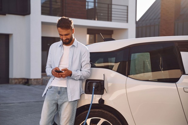 Junger Mann steht mit seinem Smartphone an sein Elektroauto gelehnt | Credit: iStock.com/standret