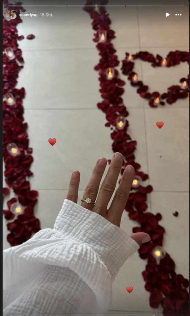 vor einem Herz mit Rosen und Kerzen zeigt Sandy ihren Ring
