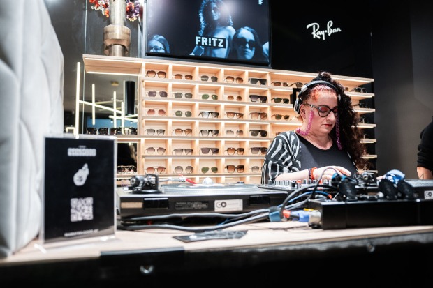 Eine Frau an einem DJ-Pult, dahinter sind Brillen zu sehen