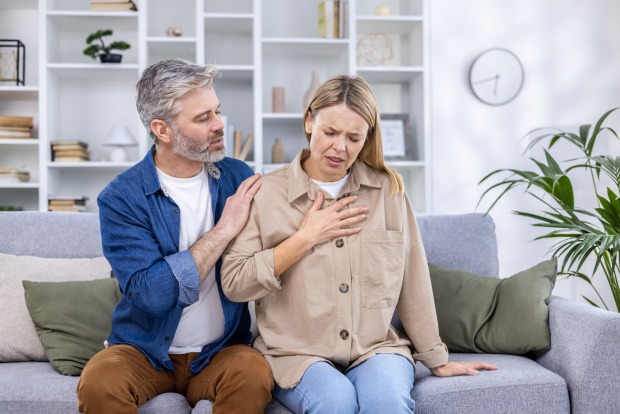 Ein Mann hält seine auf einer Couch sitzende Frau in Armen, die sich mit einem schmerzverzerrten Gesicht an die Brust greift
