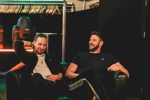 Daniel Fellner und Paul Pizzeria (Band "Aut of Orda") sitzen entspannt und lächelnd gemeinsam auf einer Couch. Im Hintergrund ist eine Stange mit vielen Kleiderbügeln zu sehen, ansonsten alles schwarz.
