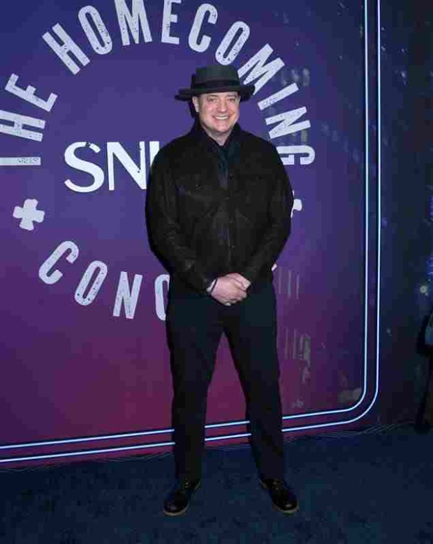 Brendan Fraser ganz in schwarz gekleidet mit Hut, lächelt in die Kamera, die Hände hat er vor seinem Körper zusammengeschlossen.