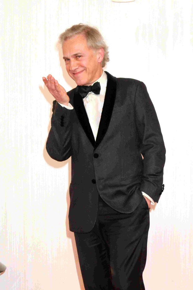 Christoph Waltz in schwarzem Smoking mit Fliege, lächelnd und eine Hand zum Kussmund geformt, vor hellem Hintergrund.
