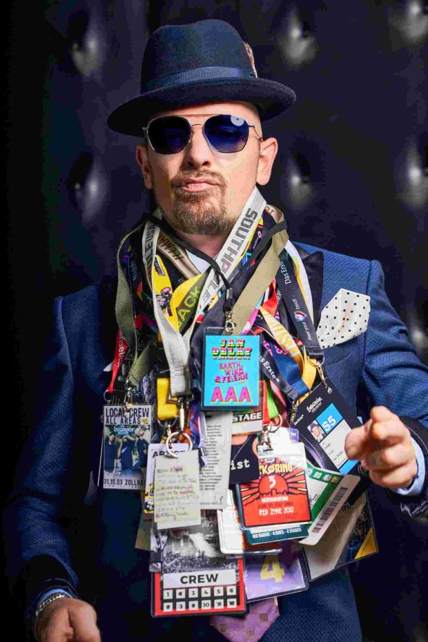 Musiker Jan Delay in dunkelblauem Sakko, Hut und Sonnenbrille. Um seinen Hals hängen mehrere Konzertpässe, sein Mund ist zu einem Duck Face geformt.