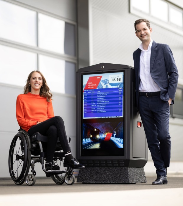 Kira Grünberg im Rollstuhl und Alexander Frank im Anzug neben einer iBox-Anzeigetafel. Die barrierefreie digitale Lösung zeigt Abfahrtszeiten und Informationen auf Augenhöhe.