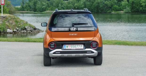 Das Heck des Hyundai Inster: Das kaffeebraune Auto parkt am Rande eines Gewässers. Am anderen Ufer sind zahlreiche Laubbäume zu sehen.