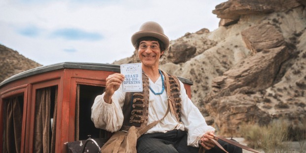 Die Figur Dimitri (Rick Kavanian) sitzt fröhlich auf einer Postkutsche in Westernkulisse und hält ein Schild mit der Aufschrift „Grand Re-Big-Opening“.