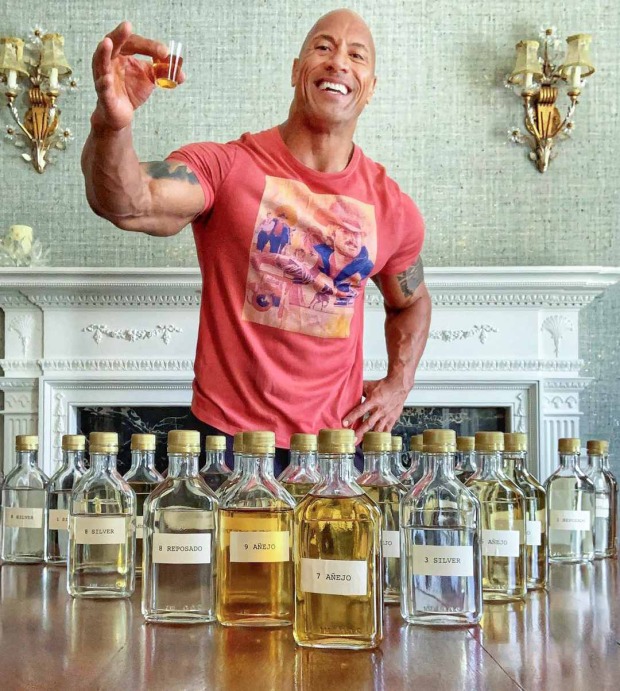 Ein muskulöser Mann in einem roten T-Shirt hält ein kleines Glas mit einer bernsteinfarbenen Flüssigkeit in die Kamera. Vor ihm stehen zahlreiche Probenflaschen mit unterschiedlichen Tequila-Sorten.
