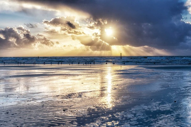 Strand auf Ameland mit spiegelndem Wasser, dramatischem Himmel und Sonnenstrahlen, die durch Wolken brechen.