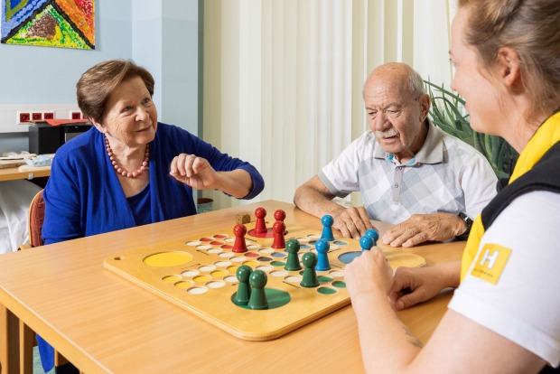 Ein Hilfswerkmitarbeiterin spielt mit zwei Senioren "Mensch ärgere dich nicht".