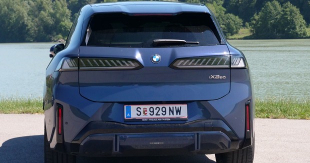 Ein hellblauer  BMW iX3 parkt am Rande eines Gewässers. Am anderen Ufer ist ein dichter Laubwald zu sehen.
