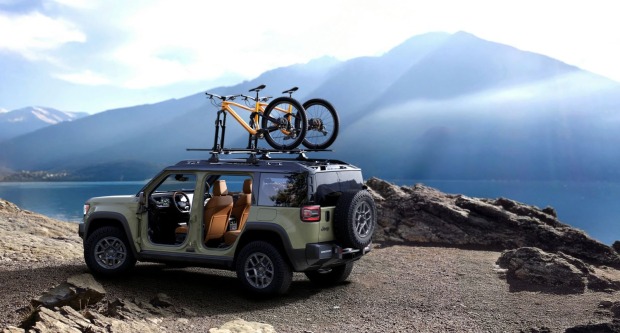 Ein olivgrüner Jeep Recon parkt auf einem felsigen Parkplatz. Auf dem Dach sind einige Fahrräder befestigt. Im Hintergrund sind ein See und Berge zu sehen.