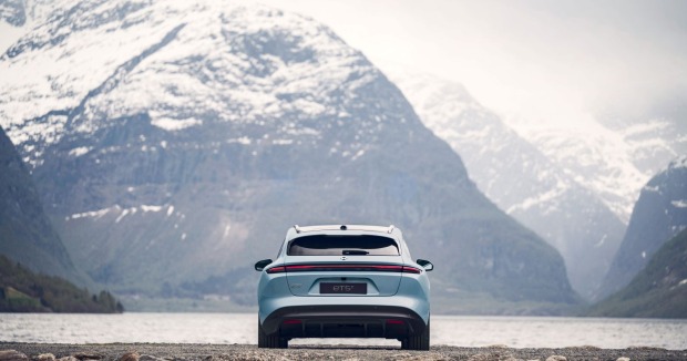 Ein hellblauer Nio ET5 Touring park vor einem See. Im Hintergrund sind hohe Berge - mit schneebedeckten Gipfeln - zu sehen.