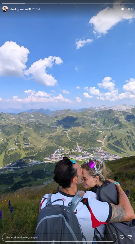 Corinna Kamper und Danilo Campisi umarmen sich in den Bergen. Danilo gibt seiner Ehefrau einen zärtlichen Kuss auf die Stirn.