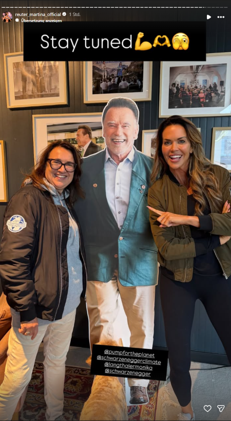 Martina Reuter posiert neben einem lebensgroßen Pappaufsteller von Arnold Schwarzenegger und kündigt auf Instagram mit „Stay tuned“ Neuigkeiten an.