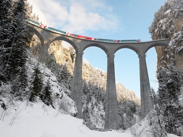 Der Glacier Express fährt über ein hohes steinernes Viadukt durch eine verschneite Wald- und Berglandschaft.