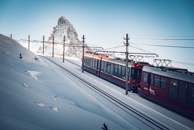 Eine rote Gornergrat Bahn fährt durch eine verschneite Hochgebirgslandschaft, im Hintergrund erhebt sich das Matterhorn.