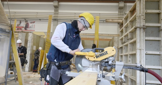 Ein junger Handwerker mit Schutzhelm und Schutzbrille arbeitet in einer Werkhalle an einer Kappsäge.