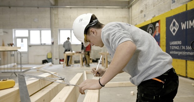 Ein junger Mann bearbeitet ein Holzstück an einer Werkbank in einer Werkhalle.
