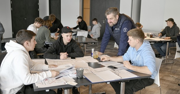 Mehrere Personen sitzen gemeinsam an einem Tisch mit Unterlagen, während ein Mann stehend etwas erklärt.