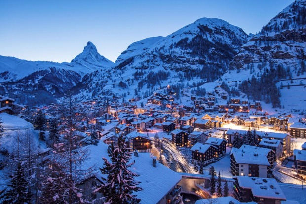 Blick auf das beleuchtete Zermatt in der Dämmerung, umgeben von verschneiten Bergen.
